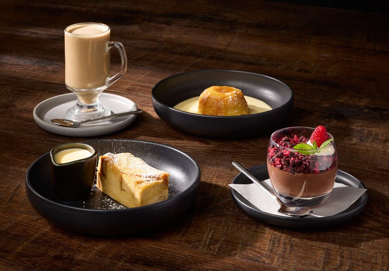 A selection of desserts including chocolate mousse topped with raspberries, a baked cheesecake served with pouring cream and a warm pudding in a pool of custard.