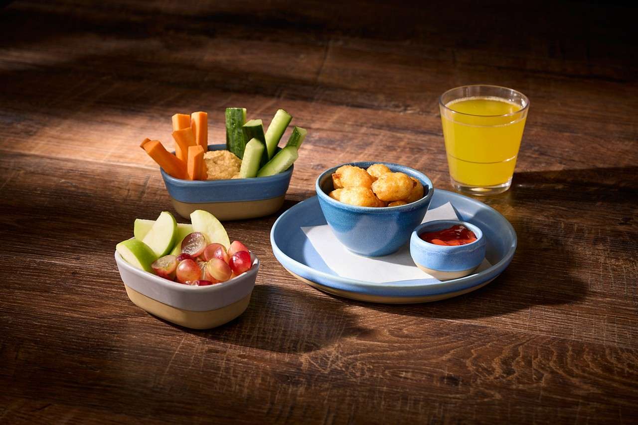 A selection of children's small bowls including hashbrown bites with a tomato dip, sliced apples and grapes and cucumber and carrot crudité.