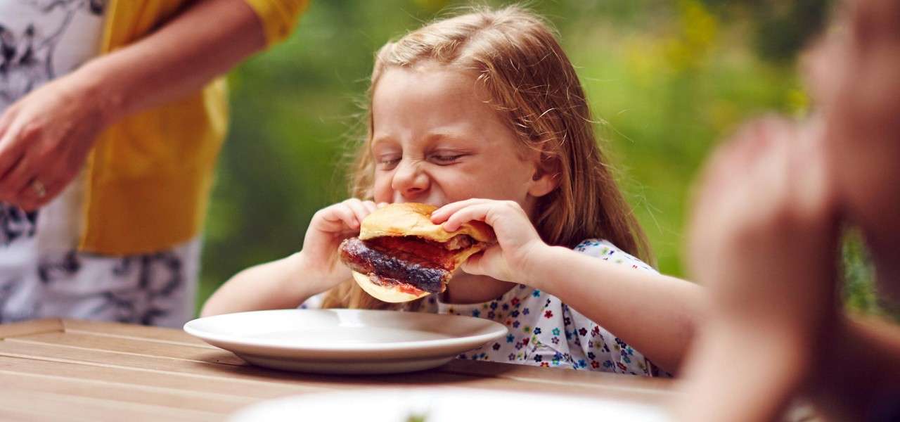 Child bites a sausage sandwich with eyes closed, gripping bun with both hands, sitting at a wooden table with empty plate, outdoors, adults nearby in background greenery.