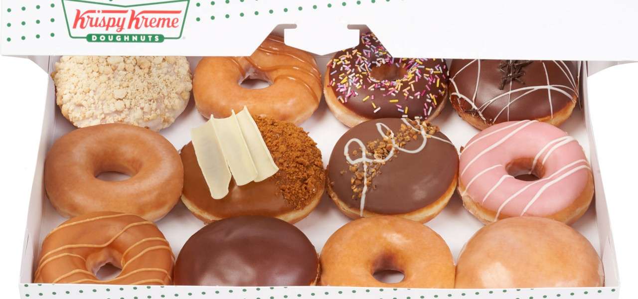 Assorted glazed and chocolate doughnuts rest in two rows, presented within an open box; context: bakery packaging with logo text “Krispy Kreme DOUGHNUTS” printed on the lid.