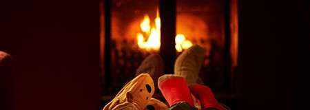 Two pairs of socked feet recline together, toes pointed toward a bright, crackling fireplace, in a dark, cozy room suggesting warmth and relaxation.