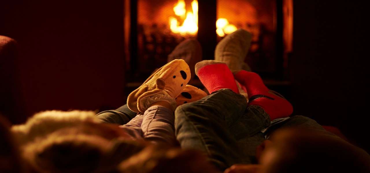 Feet in cozy socks and animal slippers rest side by side, legs outstretched, as people lounge on a couch, facing a crackling fireplace in a dim, warm living room.