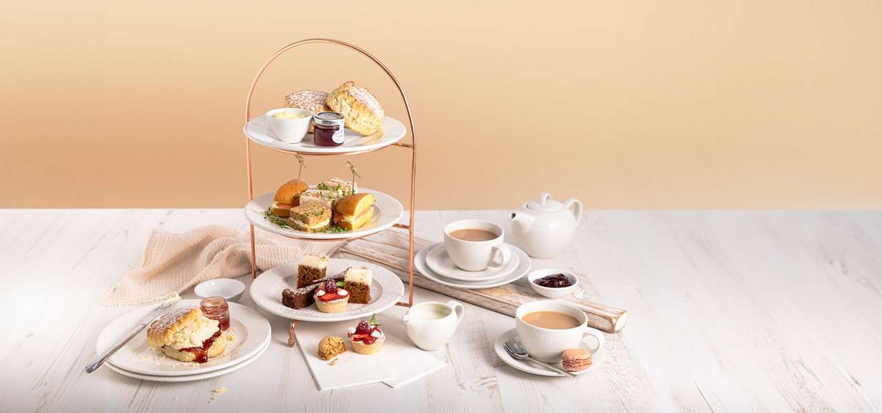 Three-tier afternoon tea stand holds scones, sandwiches, and pastries; nearby cups of tea, teapot, jam, and clotted cream sit on a light wooden table in a bright, minimal setting.