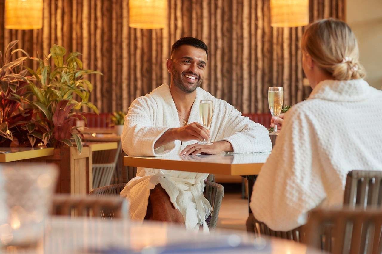 a man and women sat at a table in spa robes with glasses of prosecco