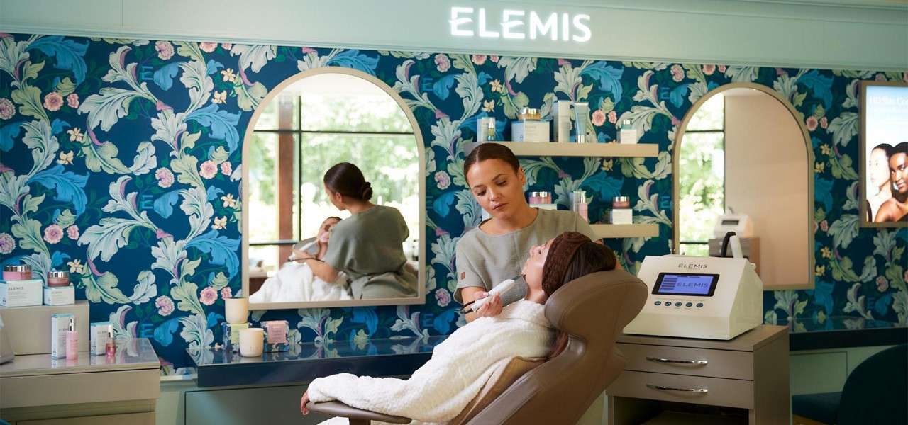 Esthetician applies a facial treatment to a reclined client, in a spa room with floral wallpaper, mirrors, and skincare shelves; illuminated signage, product labels, and a machine display read: ELEMIS.