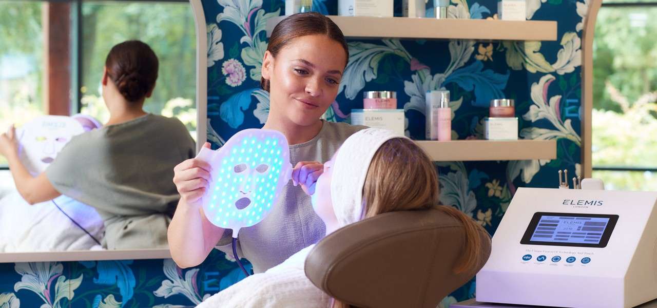 LED face mask glows blue as an esthetician holds it toward a reclining client; spa shelves and a skincare machine sit nearby. Text: ELEMIS.