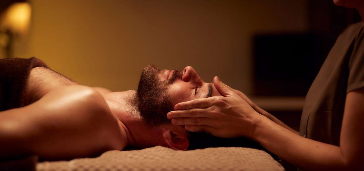 Bearded person lies on a massage table, eyes closed, receiving a gentle face massage; therapist’s hands cradle the temples in a warm, dimly lit spa room with towels.