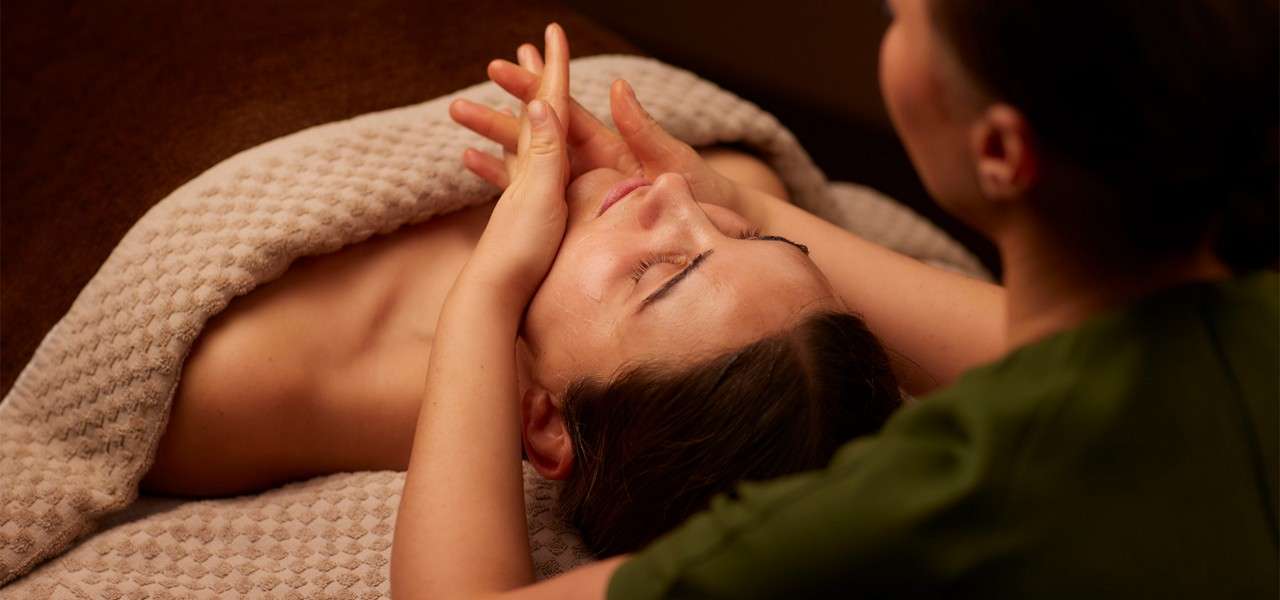 Person receives a facial massage; therapist’s hands cradle and press the cheeks and temples, while the person lies under a textured beige blanket in a warm, dimly lit spa room.