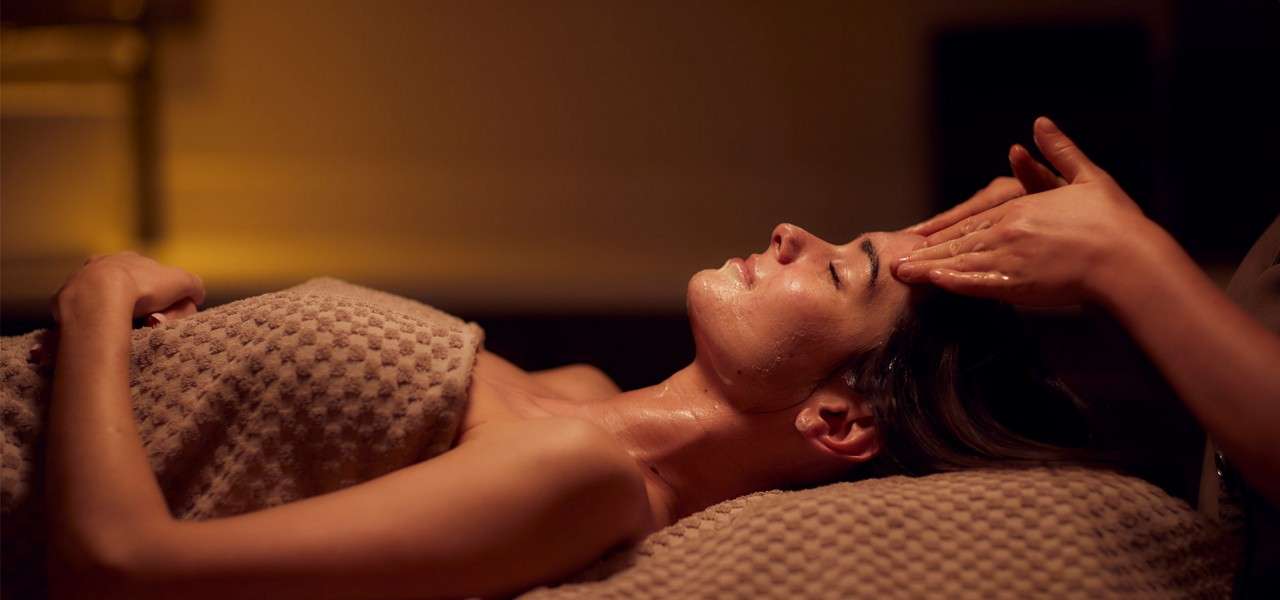 Person lies on a massage table, eyes closed, receiving an oil facial massage; therapist’s hands rub the forehead; warm ambient lighting and a textured blanket create a serene spa setting.