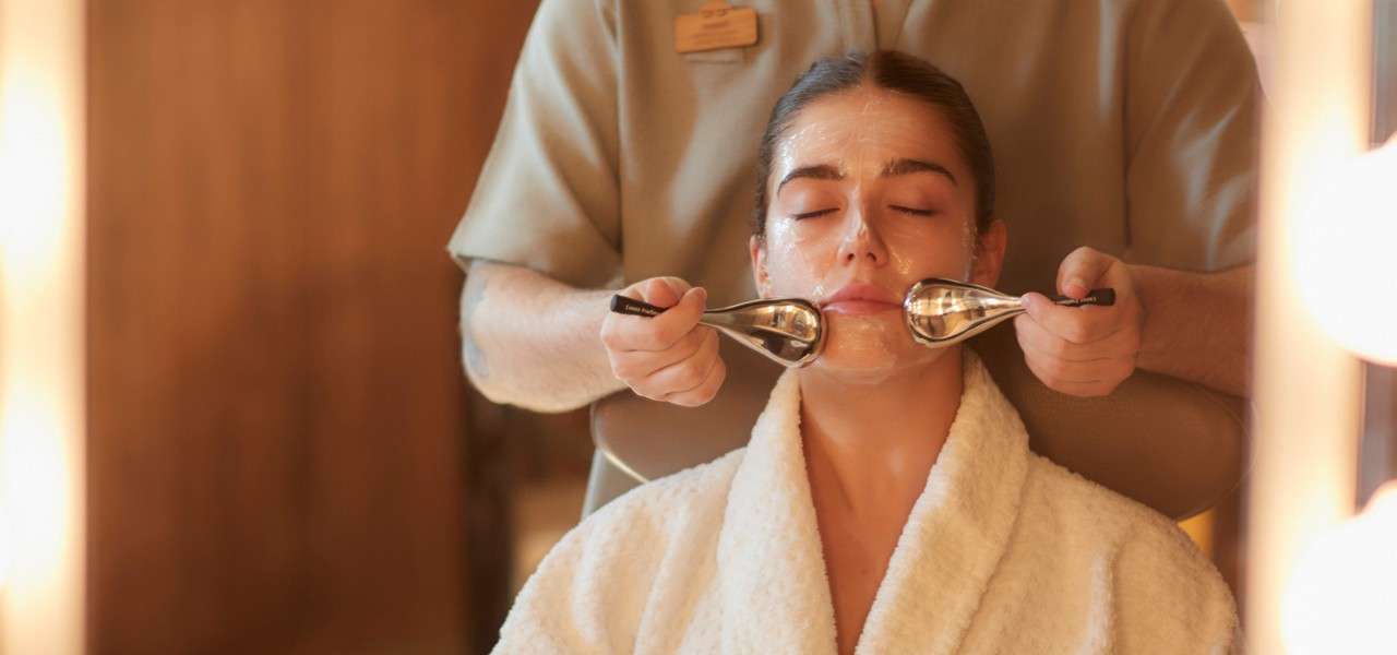 Esthetician applies chilled metal facial massagers on a client's cheeks while the client relaxes in a robe, eyes closed, inside a softly lit spa treatment room.