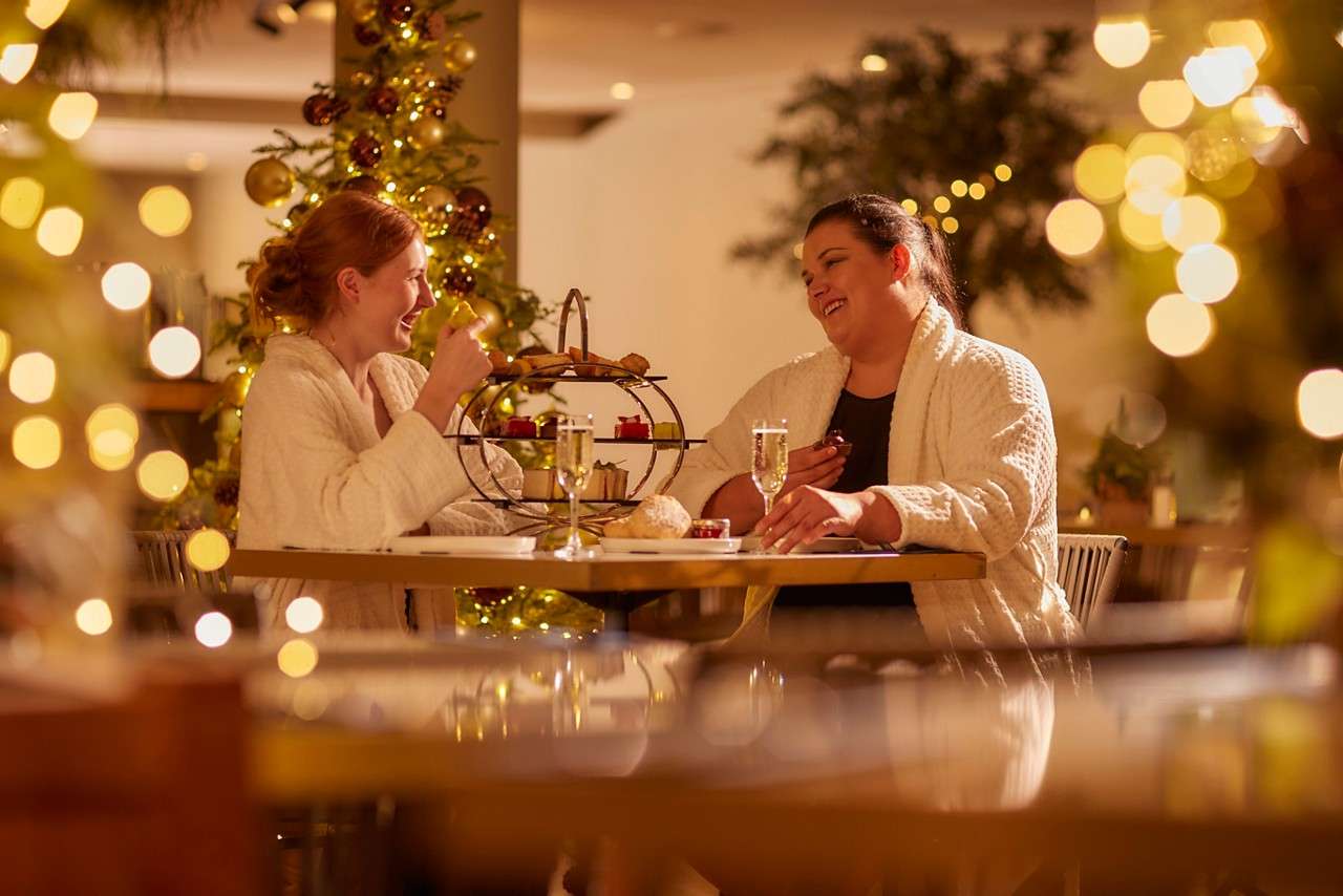 Two women in spa robes, sat at a wooden table with christmas decorations and lights around the room