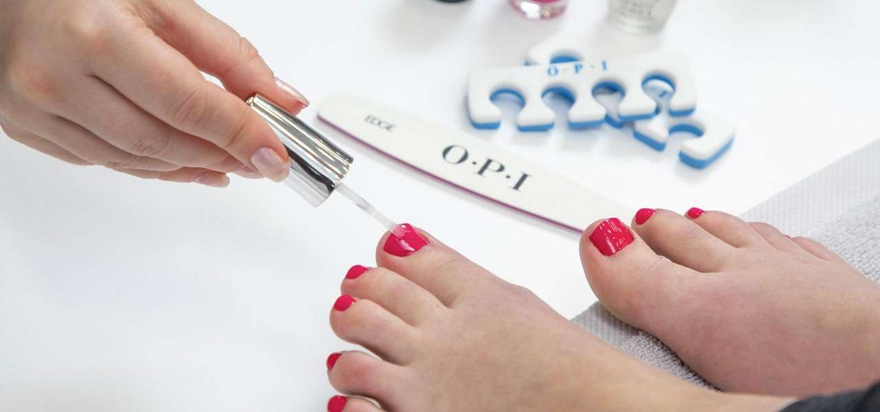 Feet receive clear polish during a pedicure; hands apply topcoat. Nearby tools include a nail file and toe separators labeled “O·P·I,” and the file also reads “EDGE.”