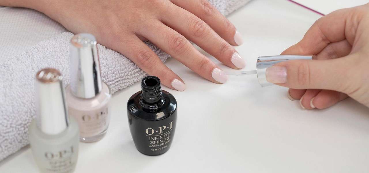 Hand with pale pink nails—another hand applies clear polish with a brush—on a salon table beside a towel and nail-polish bottles. Visible text: “O·P·I,” “INFINITE SHINE.”