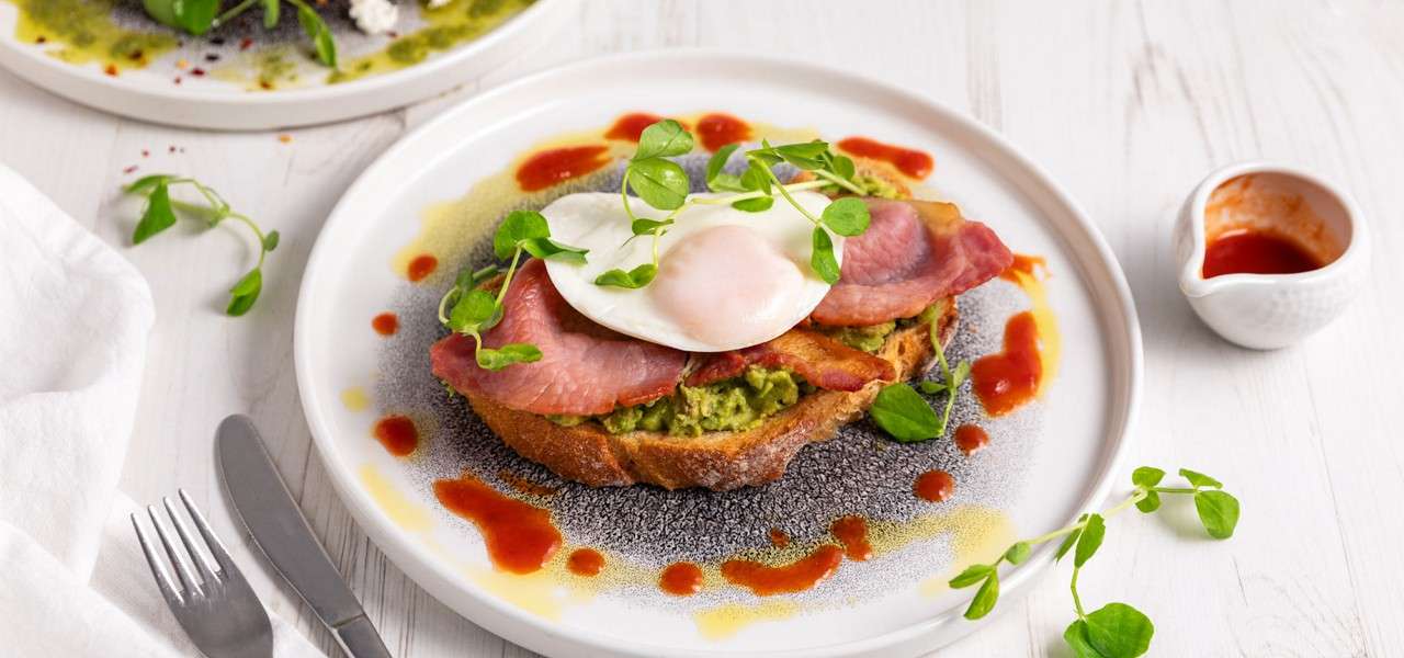 Open-faced toast carries avocado mash, bacon, and a poached egg, garnished with microgreens; drizzled sauces surround it on a white plate beside cutlery and a sauce jug on a table.