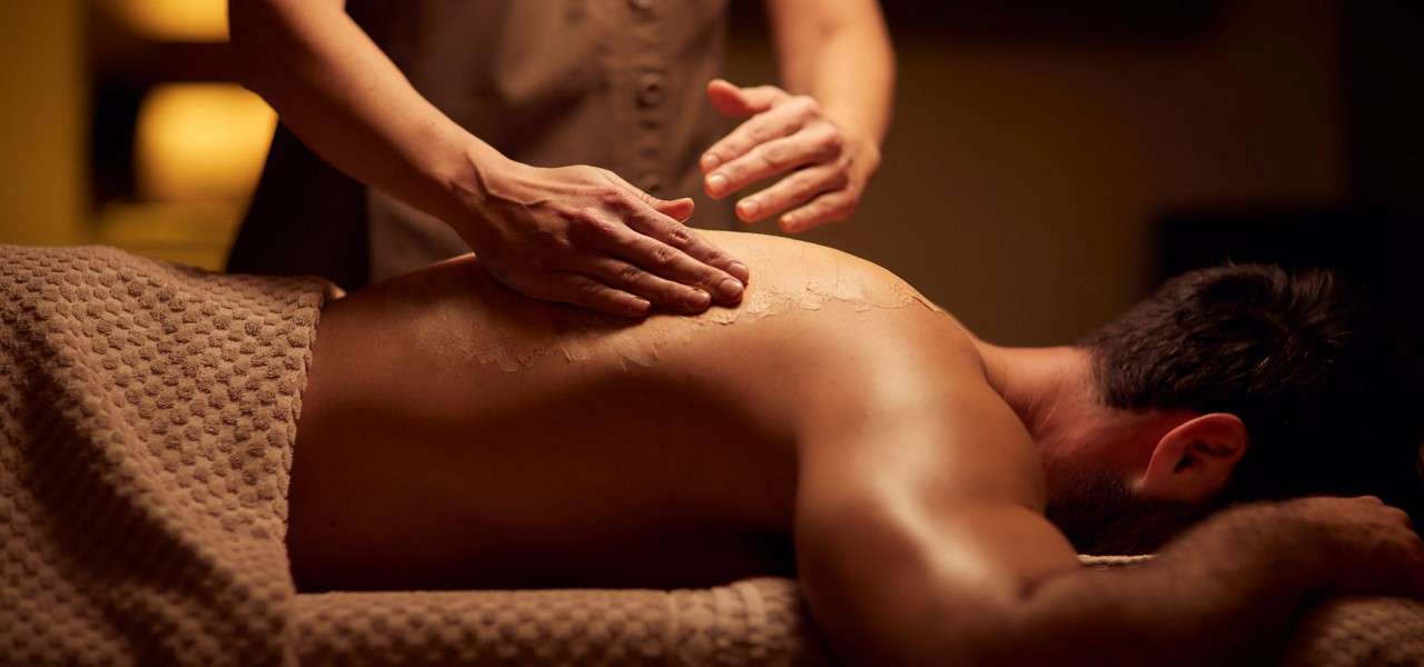Massage therapist’s hands spread lotion across a man’s bare back, kneading muscles. He lies face-down on a treatment table under a textured towel, in warm, dim spa lighting.