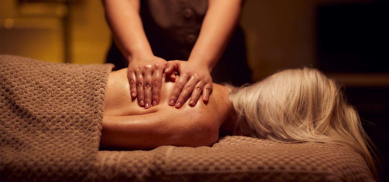 Oiled hands press and knead a person’s bare back, action: massage. Context: recipient lies face-down on a massage table under a textured brown towel in warm, dim spa lighting.