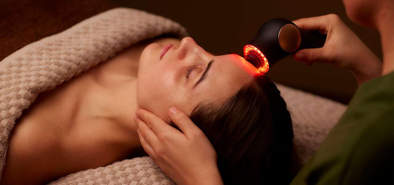 Handheld red-light device treats a person's forehead, while a practitioner supports their head, in a calm spa setting with warm lighting and a textured beige blanket.