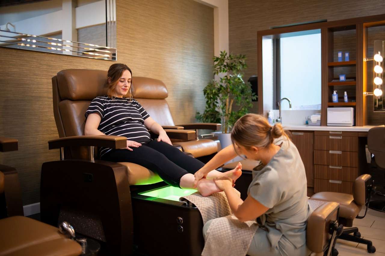Client reclines on a pedicure chair, receiving a foot massage from a technician, in a modern spa with brown leather seats, glowing green footbath, shelves, sink, and vanity lights.