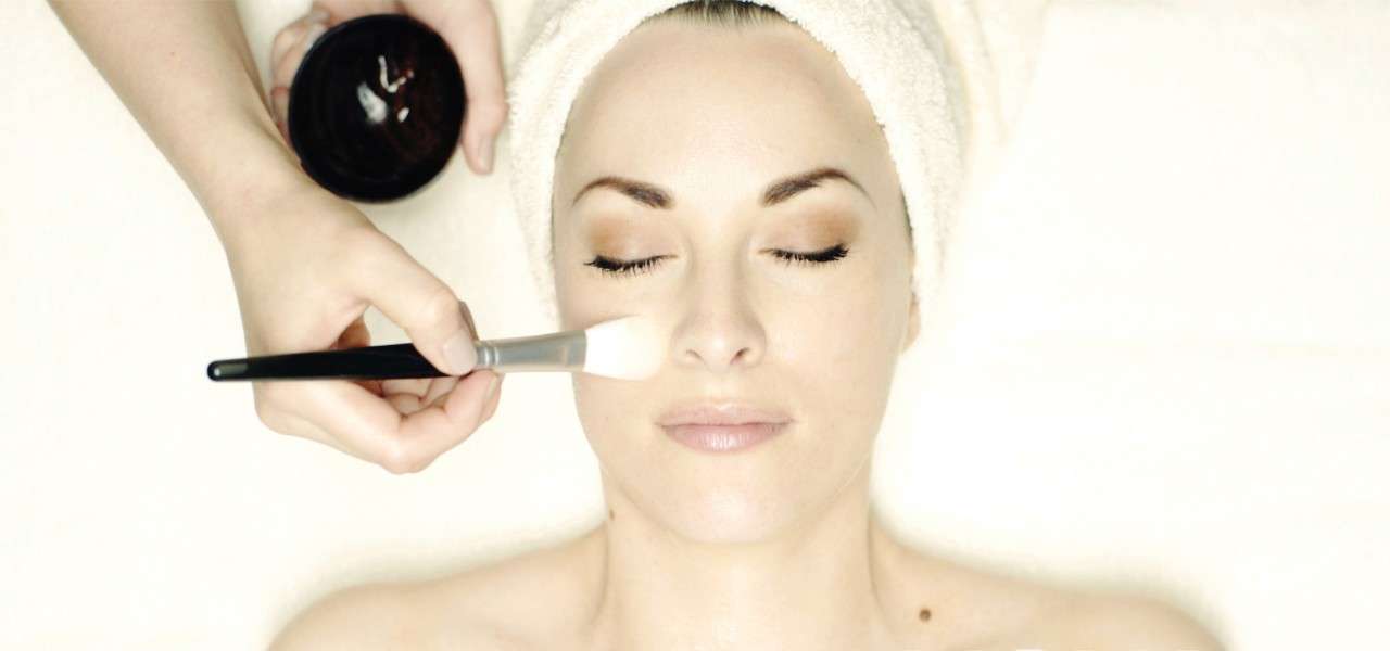 A woman laying with her eyes closed whilst a cream is applied to her face with a brush