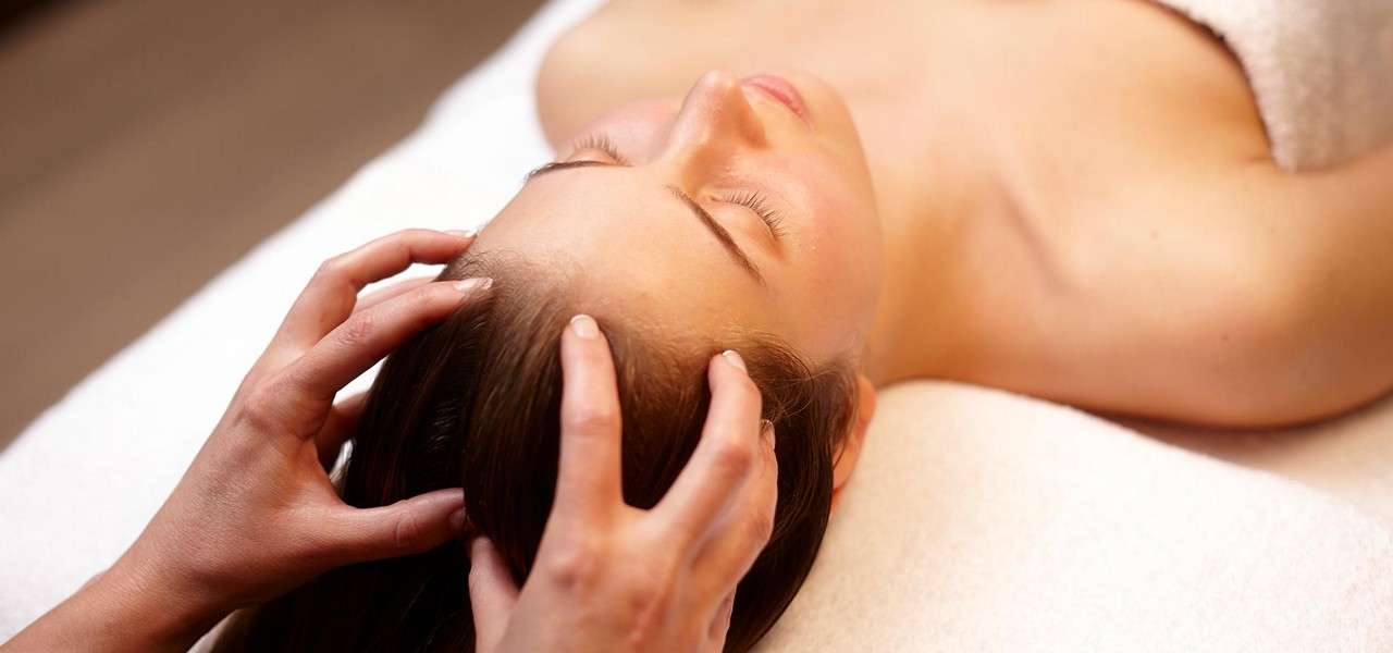 Client receives a scalp massage; hands press and knead the hairline as the person lies on a towel-covered table in a calm spa setting.