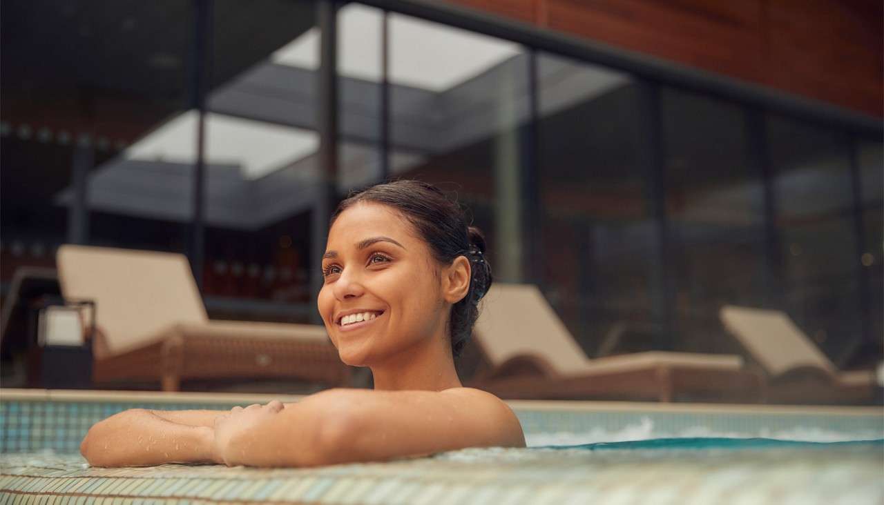 Swimming pool reflects sunlight, water calm; ladders at corners. Surrounded by a two-story colonnade with glass walls, hanging green vines, and potted plants, forming a serene courtyard enclosure.