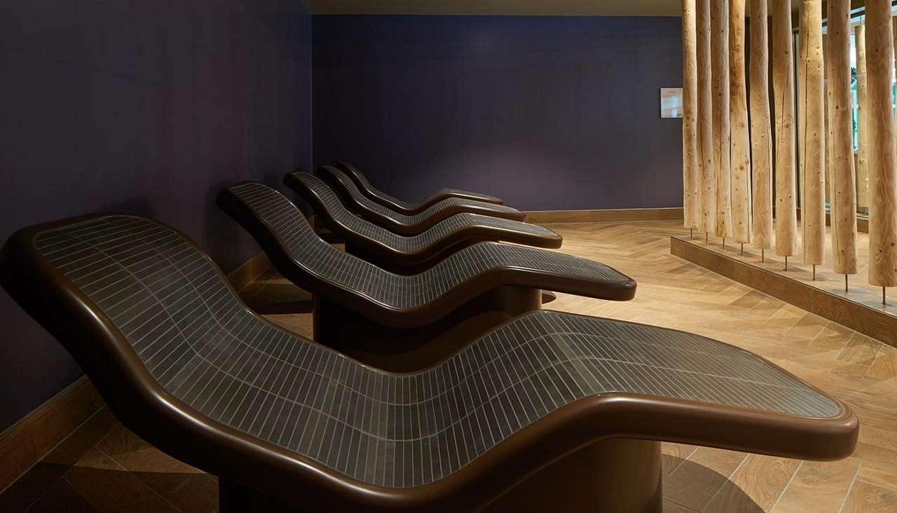 Circular cushioned bench curves around the room, glowing with red uplighting, beneath an oval skylight depicting blue sky and leaves, surrounded by tall blue curtains in a dim, modern lounge.