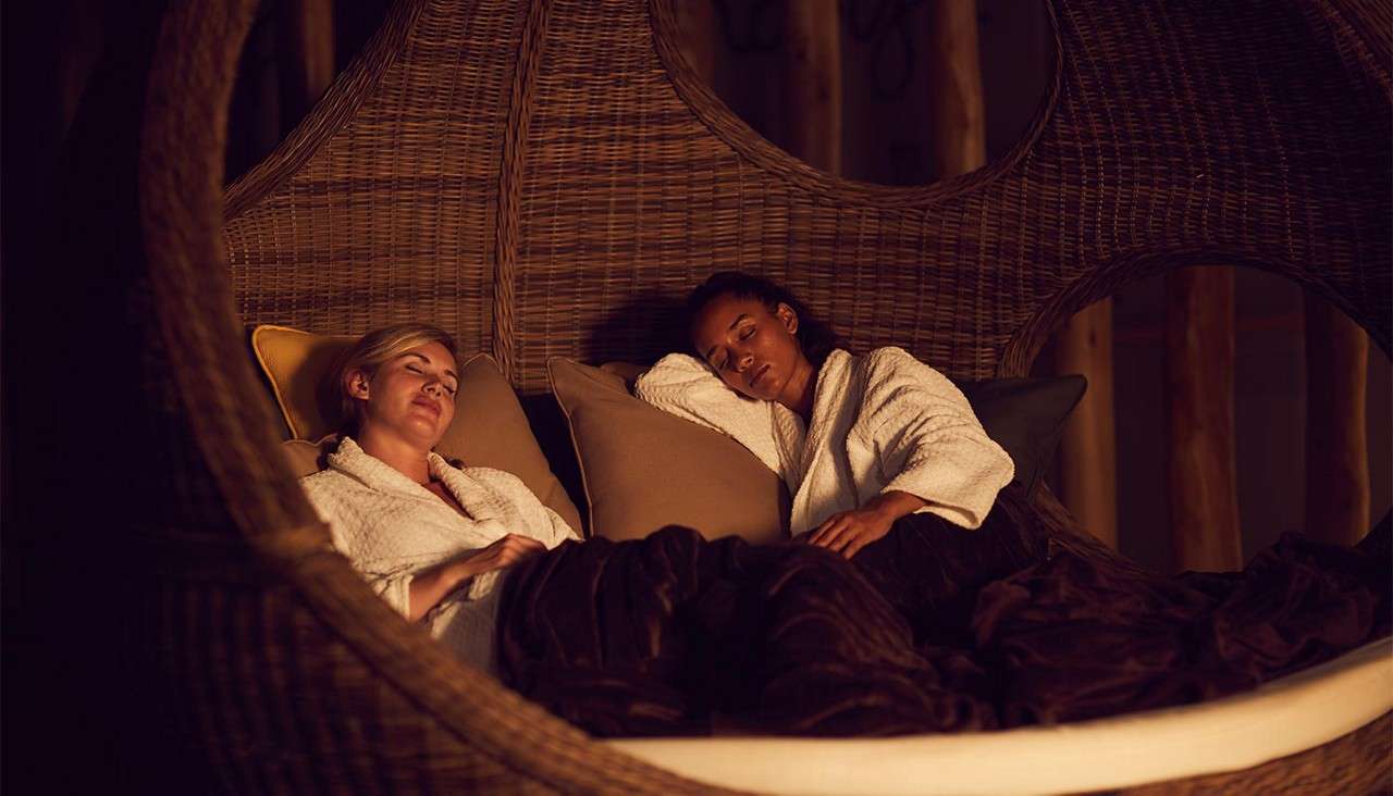 Person in a black swimsuit sits with knees hugged, relaxing on a wooden bench, gazing out a large window; in a wooden sauna with wall thermometer, overlooking lush green trees.