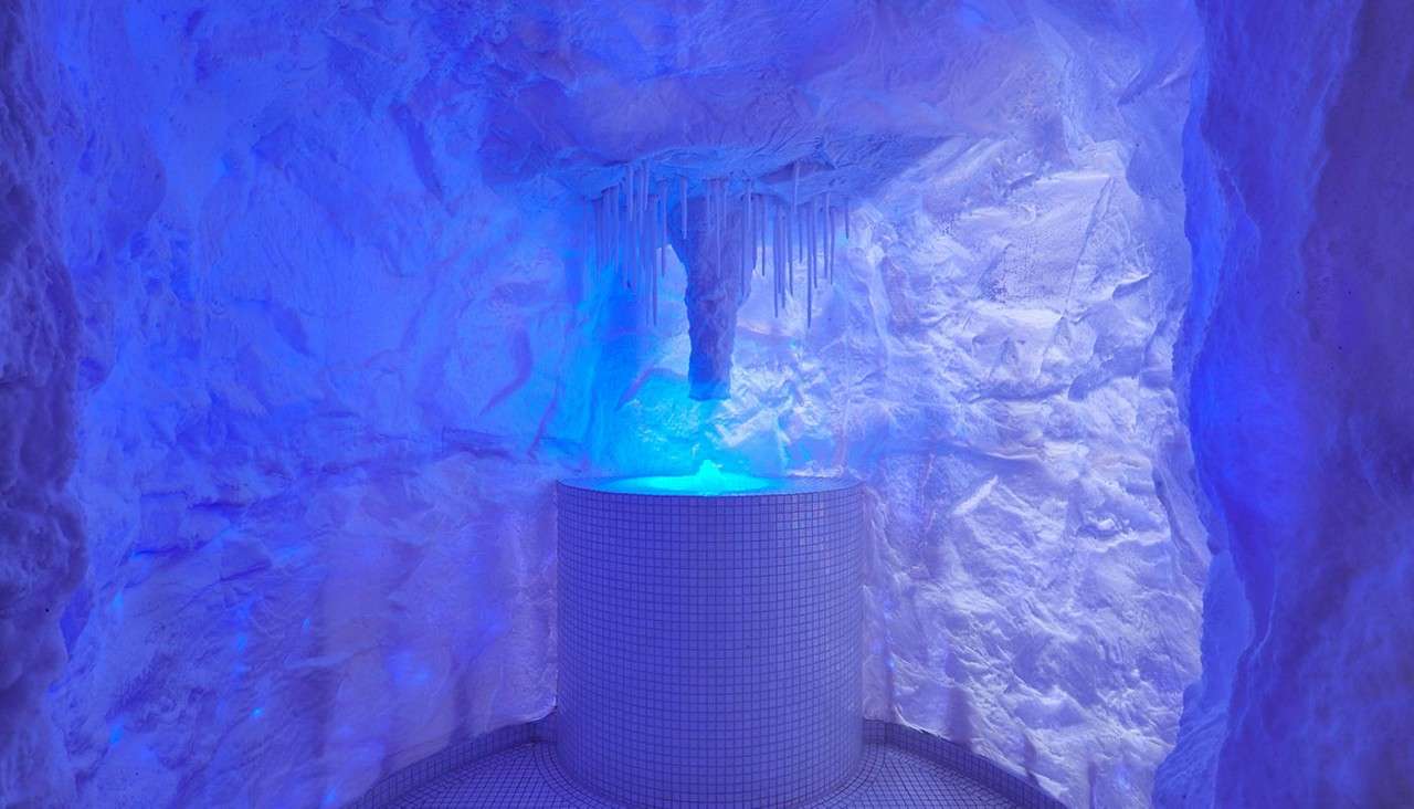 Fire pit table glows warmly, casting light on a cushioned bench. Inside a wood-paneled cabin, pendant lamps hang overhead; fur throws and animal hides decorate walls, with a forest view.