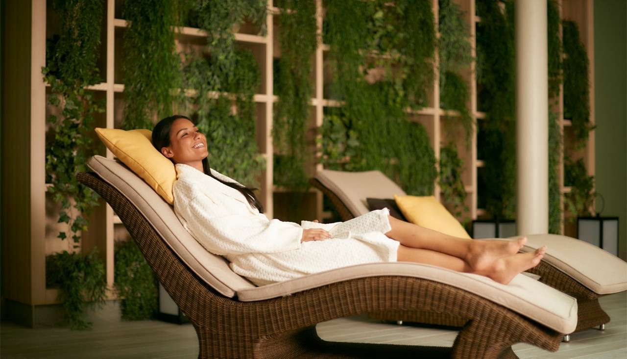 Two people in white robes recline and doze, wrapped in dark blankets, on a large woven rattan pod seat with cushions, set in a dim, warm-lit lounge with timber columns.