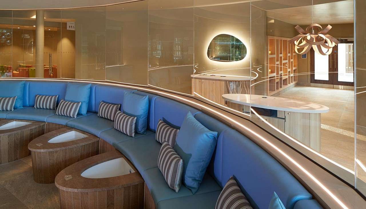 Stone basins stand clustered, trickling water, within a circular screen of vertical timber poles; behind them, floor-to-ceiling windows reveal an outdoor pool and trees in a serene spa.