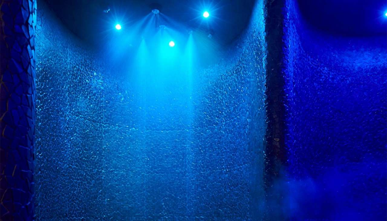 Wicker pod chairs cradle cushions, arranged side by side, in a dim lounge. Soft spotlights glow above, wood floor beneath, and a glass wall with curtains borders the seating.