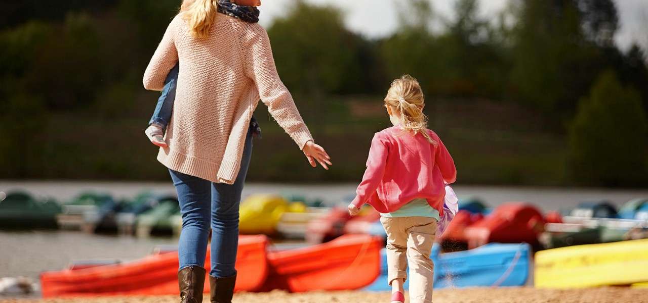 Adult and child walk on a sandy shore toward colorful kayaks; the adult in a sweater and boots reaches toward the running child, with calm water and tree-lined background.