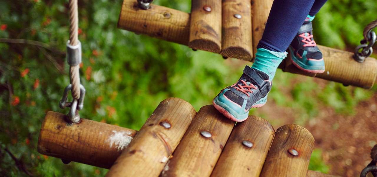 Child’s sneakered feet step cautiously across a suspended wooden log bridge, connected by chains and cables, within a forest ropes course.