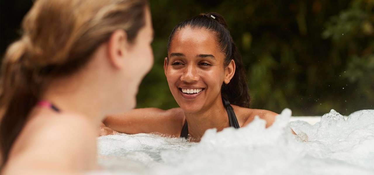 Two people converse while relaxing in a bubbling hot tub, one smiling at the other. Foamy water churns in the foreground; leafy greenery forms a soft, outdoor background.