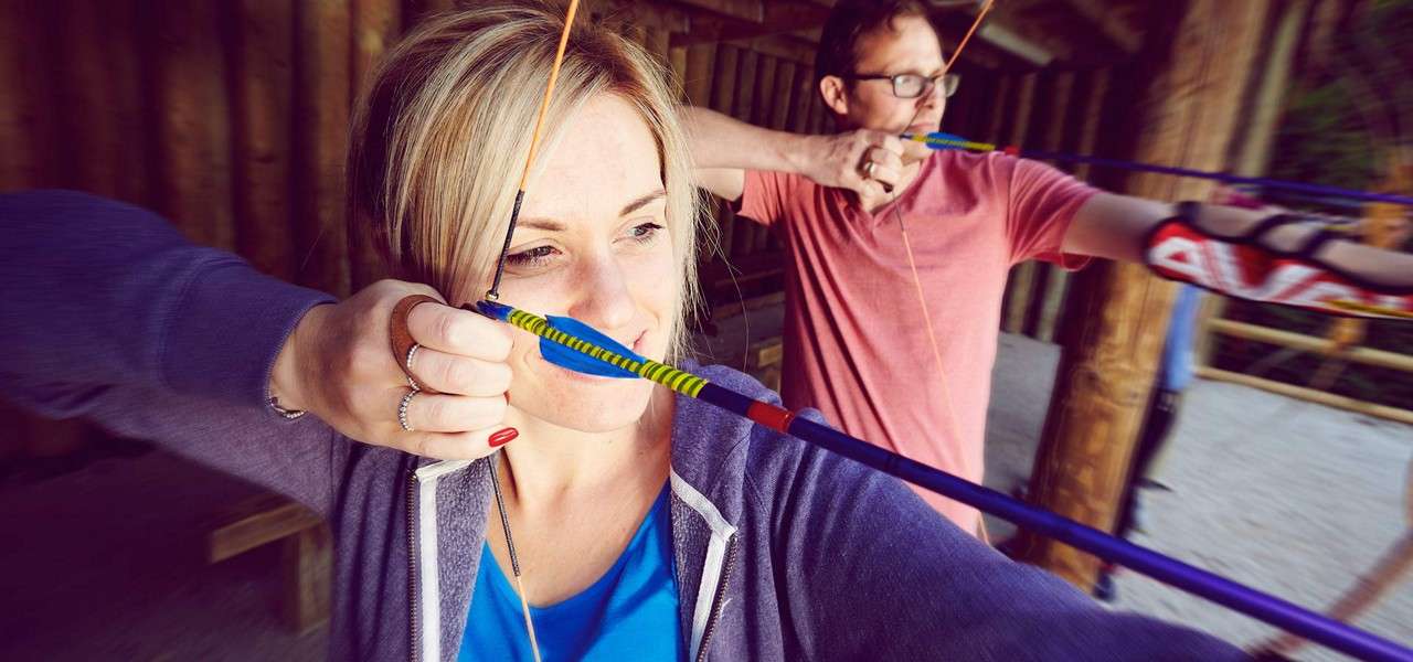 Two archers draw recurve bows, arrows nocked near their cheeks, aiming downrange under a wooden shelter. The foreground woman focuses; the man behind wears glasses and an armguard.