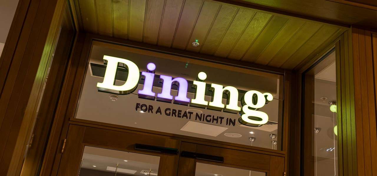 Illuminated “Dining” sign glows above a wooden-framed glass entrance; below, smaller text reads: “FOR A GREAT NIGHT IN.” Warm interior lights reflect inside.