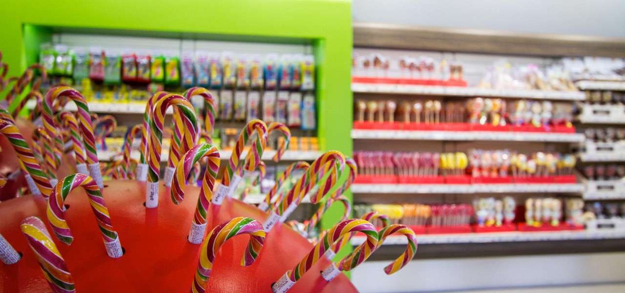 Colorful candy canes stand upright in a display holder, inviting selection, within a brightly lit candy shop filled with shelves of lollipops and packaged sweets in vivid colors.