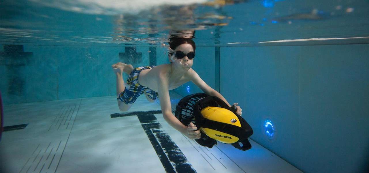 Child wearing goggles rides a yellow underwater scooter labeled “SEA-DOO,” gliding forward and holding handles. Context: clear swimming pool, tiled floor, lane markings, and blue wall lights.