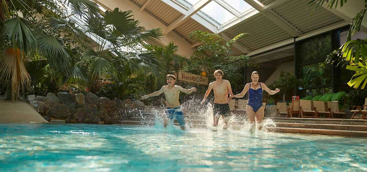 Three swimmers run forward, splashing into a turquoise indoor pool at a tropical-themed waterpark, surrounded by palm plants, lounge chairs, and skylights. Visible sign: VENTURE BAY.