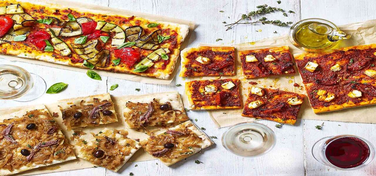 Rectangular pizzas sit sliced on parchment, topped with grilled vegetables, cheese in tomato sauce, and caramelized onions with anchovies. Arranged on a wooden table with basil, olive oil, and wine.