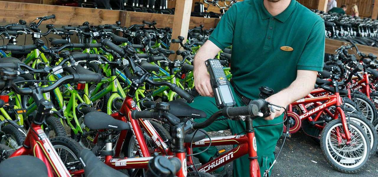 Worker scans a red children’s bicycle with a handheld device, surrounded by rows of red and green bikes in an outdoor covered rental area. Text: “Schwinn,” “HIRE BIKE.”