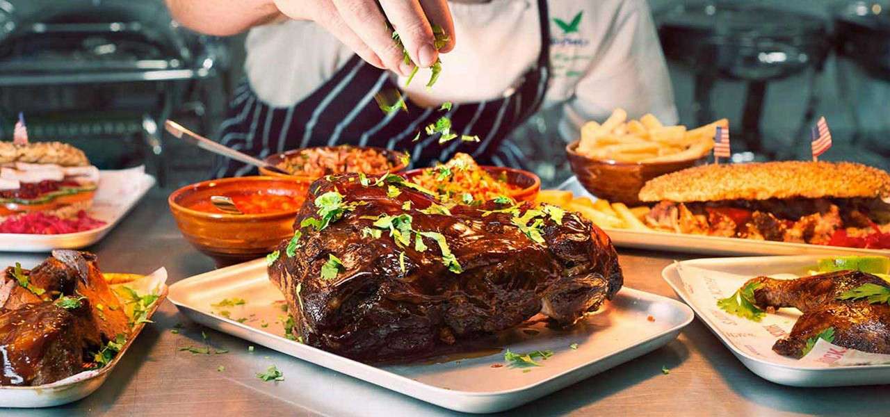 Glazed roasted meat sits on a tray as a chef sprinkles fresh herbs, surrounded by bowls of rice and sauce, fries, sandwiches, and grilled chicken on a stainless kitchen counter.