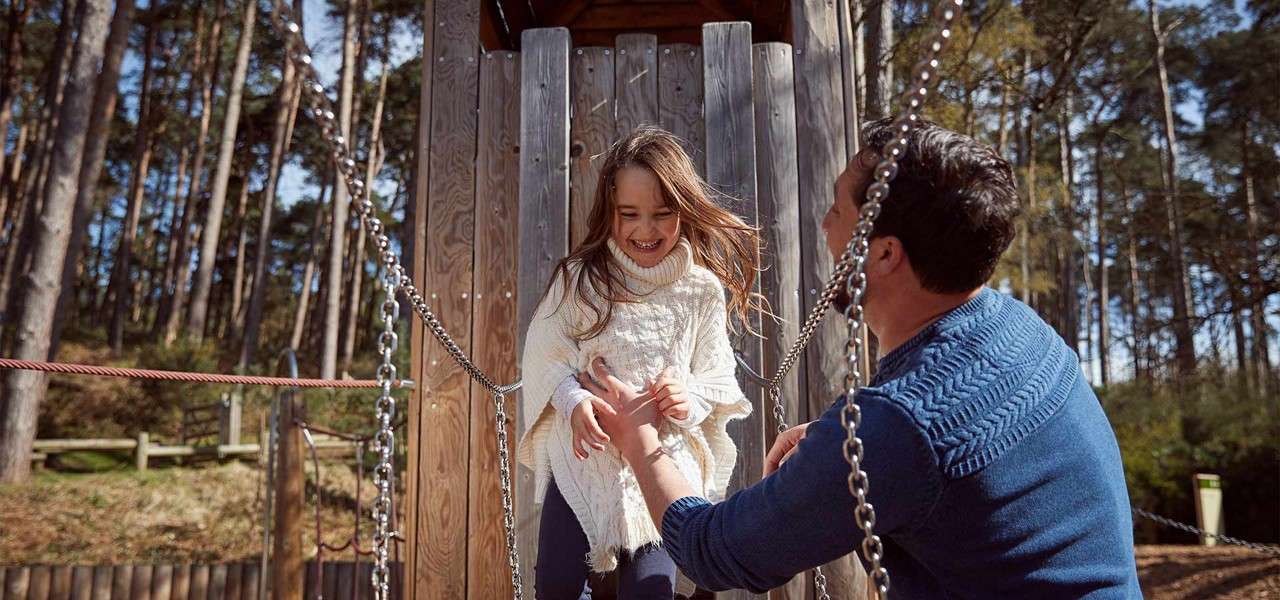 Child in white sweater descends a wooden play tower, laughing as an adult in blue reaches to catch her, amid chain railings, ropes, and tall pines in a forested playground.