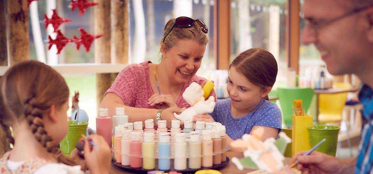 Children and adults paint ceramic figurines, selecting colors from a rotating tray of pastel paint bottles, in a bright craft studio with large windows, green chairs, and red hanging decorations.