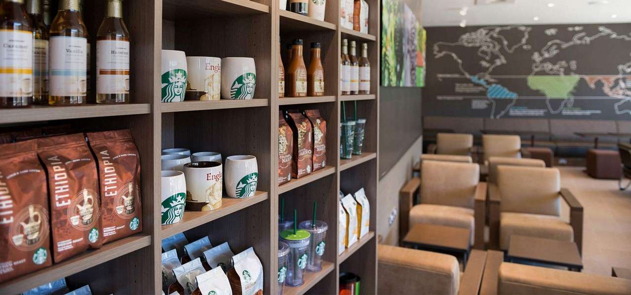 Shelves display coffee bags, syrups, mugs, and tumblers; seating extends beyond in a modern café with a world-map wall. Visible text: Caramel, Vanilla, Hazelnut, ETHIOPIA.