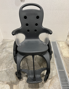 Pediatric-style shower/commode wheelchair stands unused, featuring a molded plastic seat with drainage holes, armrests, split footrest, and large rear wheels, positioned on a tiled floor beside a metal floor drain.