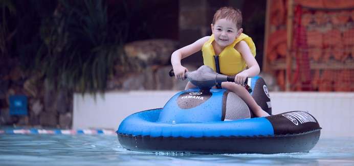 Child rides a blue inflatable bumper boat, gripping handlebars and smiling, gliding across a shallow pool, with greenery and poolside structures in the background.