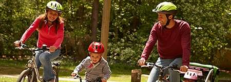 Two adults and a child ride bicycles wearing helmets along a park path; trees and grass surround them, and a bike trailer with a pink helmet sits behind one bicycle.