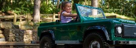 Child steers a green mini off-road vehicle labeled "DEFENDER" along a path, smiling; wooded park surrounds with trees, wooden fence, and stone wall.