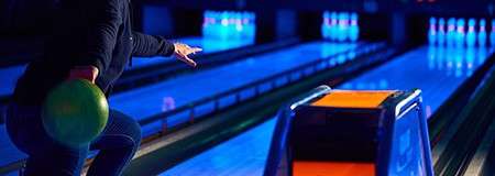 Bowling ball rolls from a bowler’s hand toward illuminated pins under neon-blue lighting. Lanes stretch ahead with gutters and a ball ramp visible, creating a glowing, arcade-like alley atmosphere.